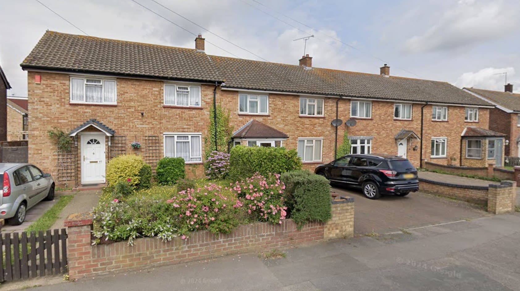 Claudian Way, Chadwell St Mary: brown-brick terraced and semi-detached two-storey homes along the street with front gardens, hedges and parked cars, Thurrock Council scheme