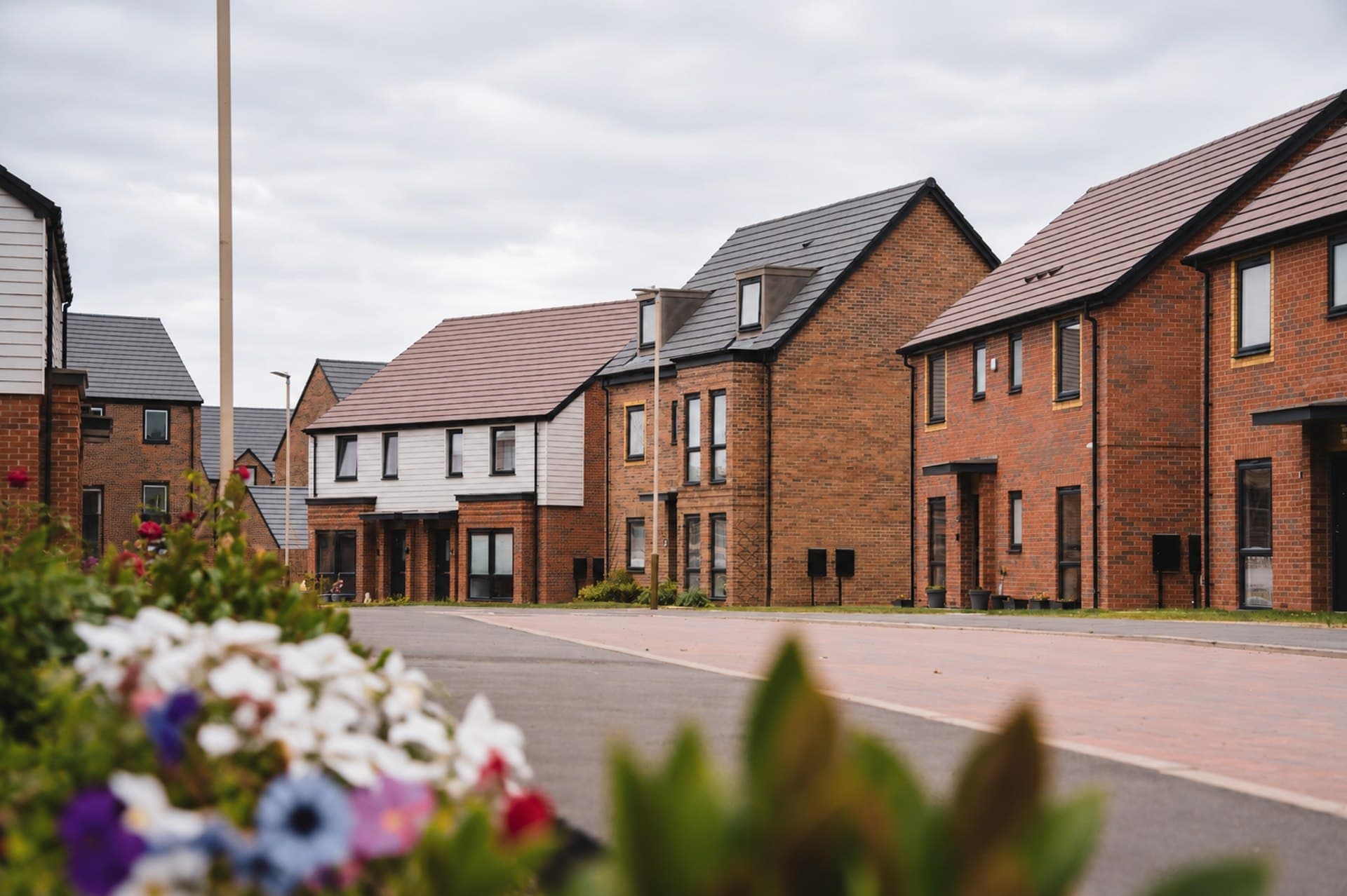 Leicester New Build, Leicester: street view of completed red-brick two-storey homes with white-render accents and anthracite tiled roofs along a new street, a flowering verge in the foreground, Partner Construction / Balfour Beatty scheme