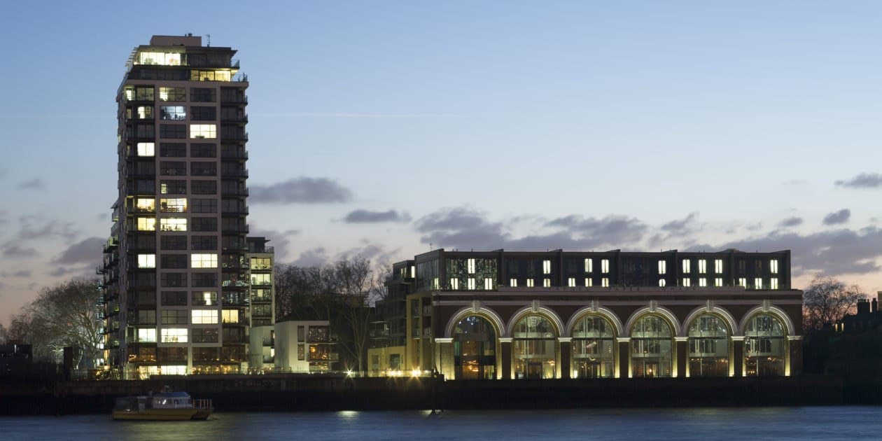 Paynes & Borthwick Wharves, West Greenwich: riverside view at dusk across the Thames, the illuminated residential tower beside the restored brick wharf building, lights reflecting on the water