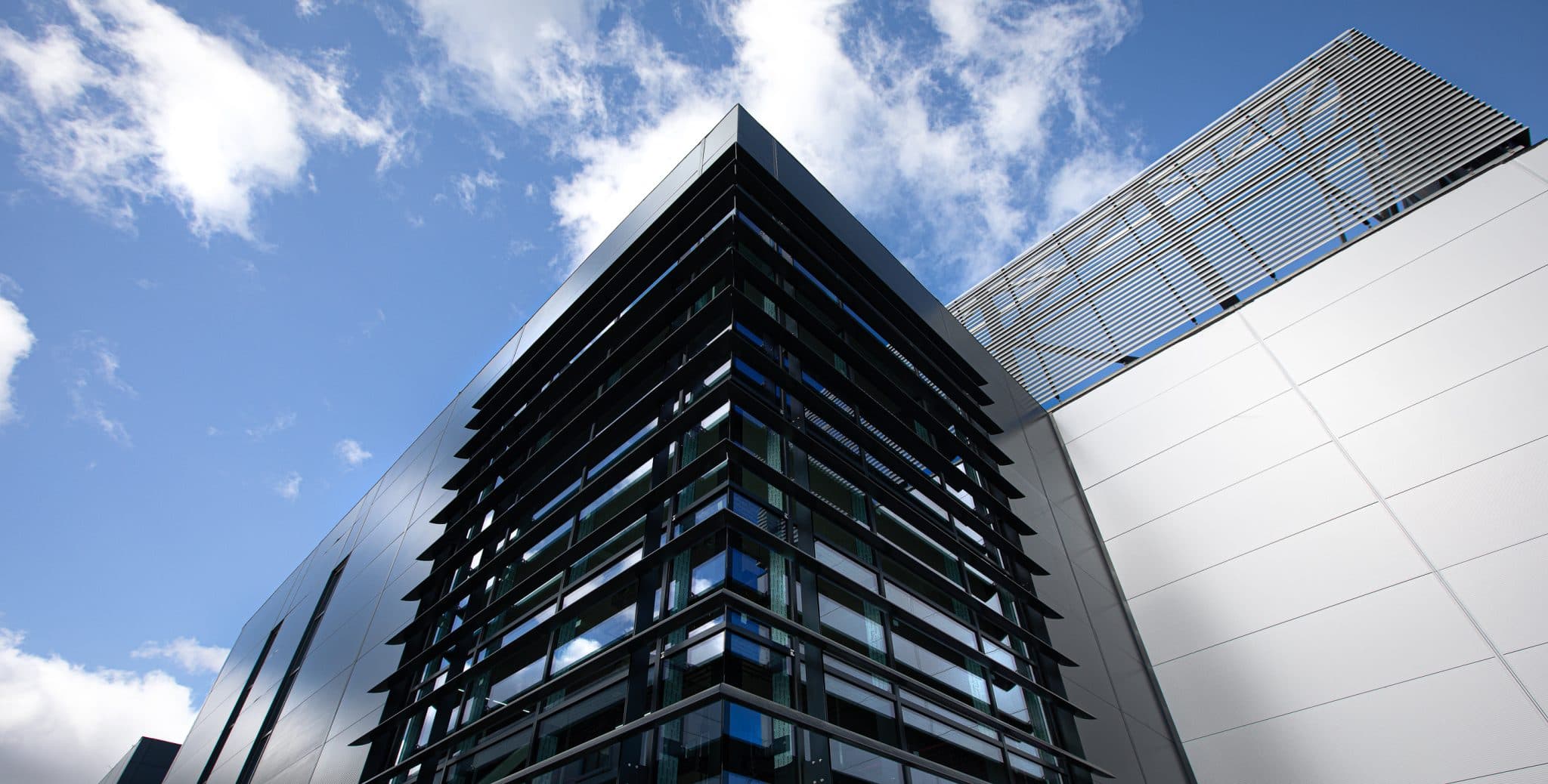 Points Cross Phase 1 (Blocks A and B), Leeds South Bank: corner view looking up at a contemporary mixed-tenure block clad in angled black glazing and aluminium panels, framed against a clear blue sky with white clouds, Guinness Partnership shared-ownership scheme.