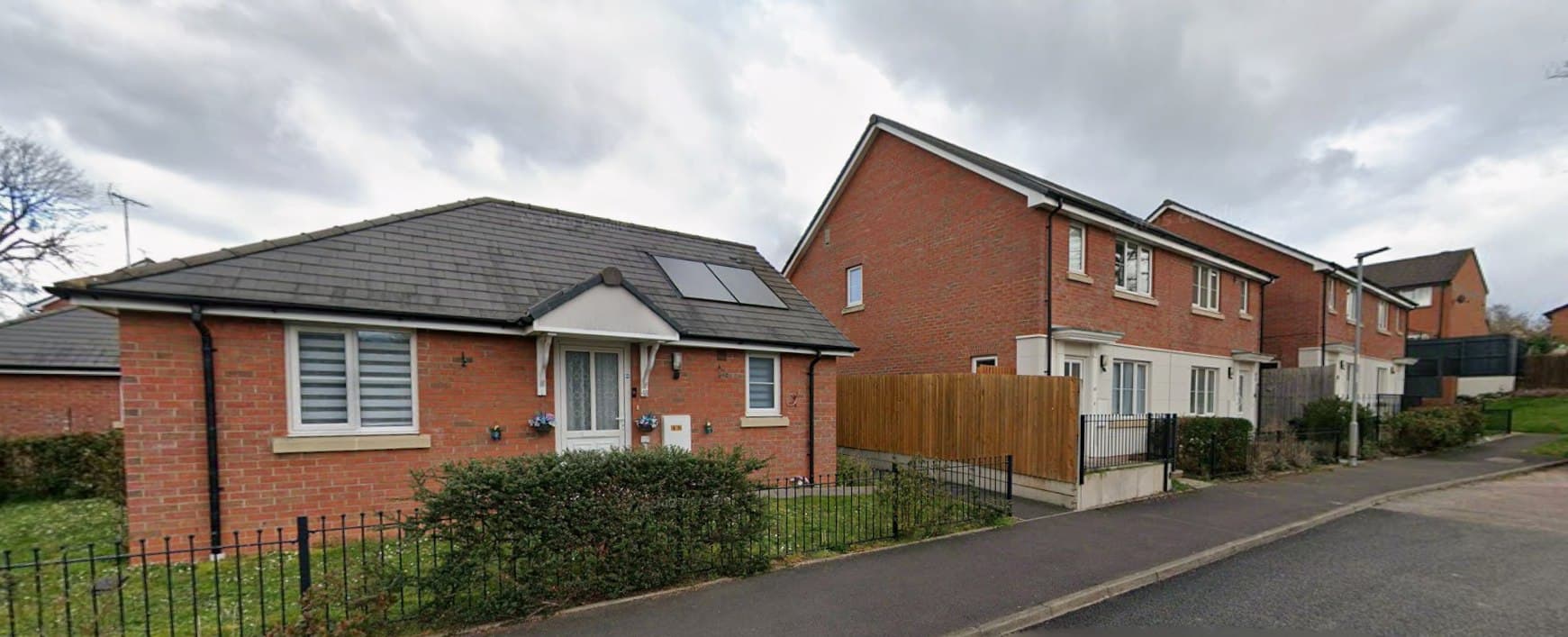 Smallbrook Gardens, Ross-on-Wye: single-storey red-brick bungalow with roof-mounted solar panels alongside two-storey brick homes behind railings, WM Housing scheme