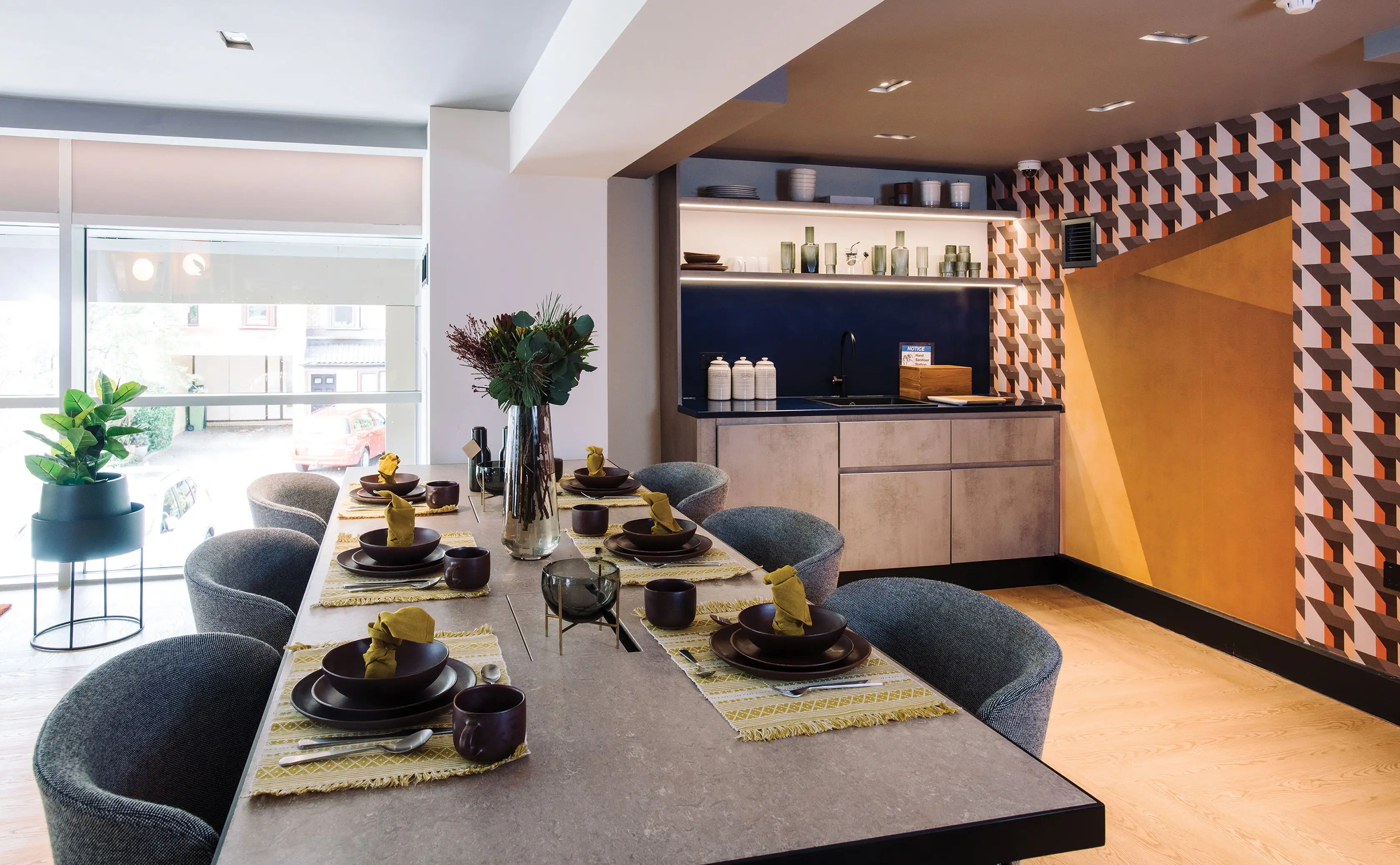 Solstice Apartments, Milton Keynes: close-up of the shared dining table dressed for service, kitchenette and geometric orange-and-black wall feature beyond