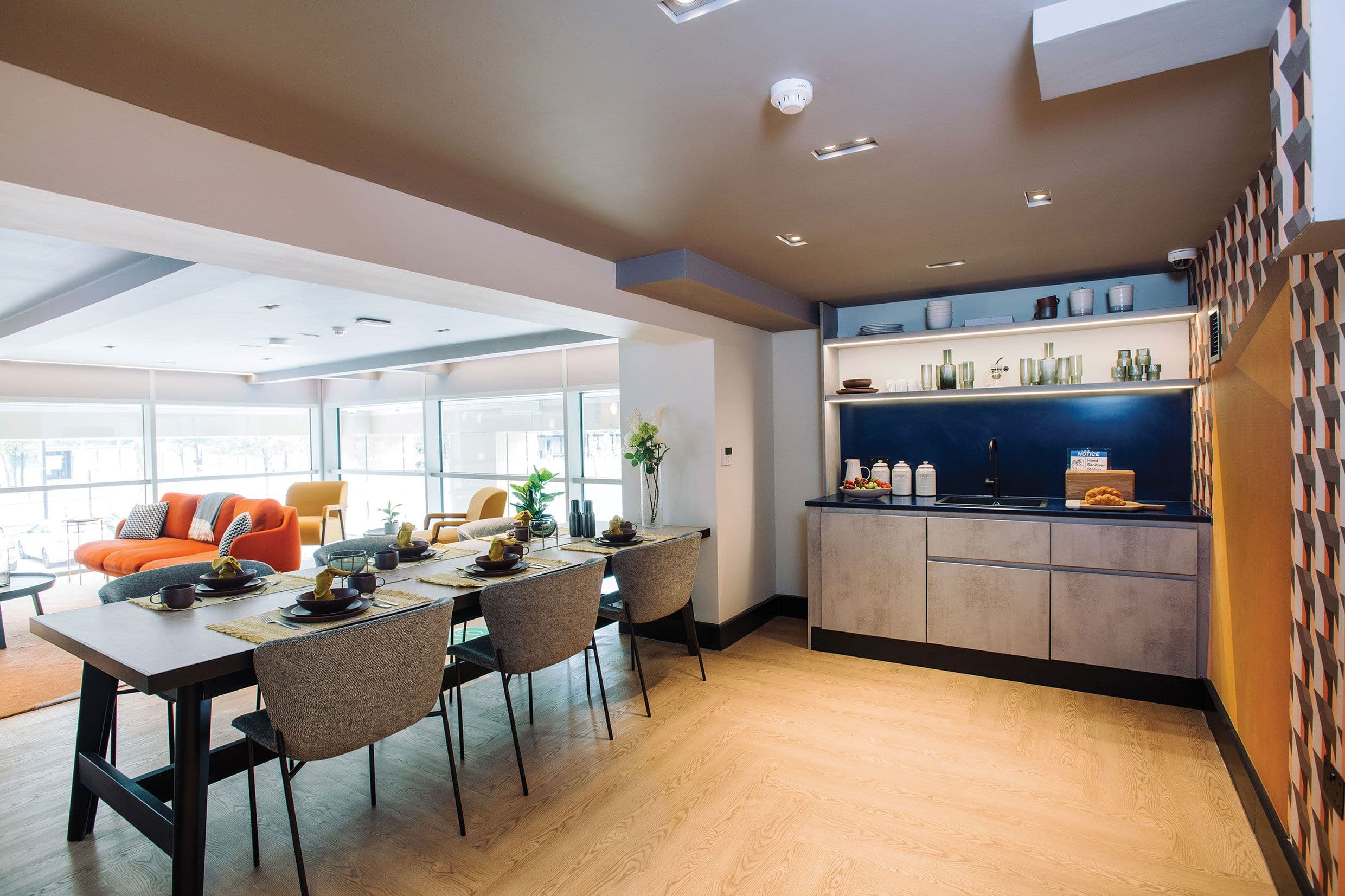 Solstice Apartments, Milton Keynes: shared dining and lounge area with long table, fabric chairs and kitchenette under a navy backsplash, full-height glazing along one side