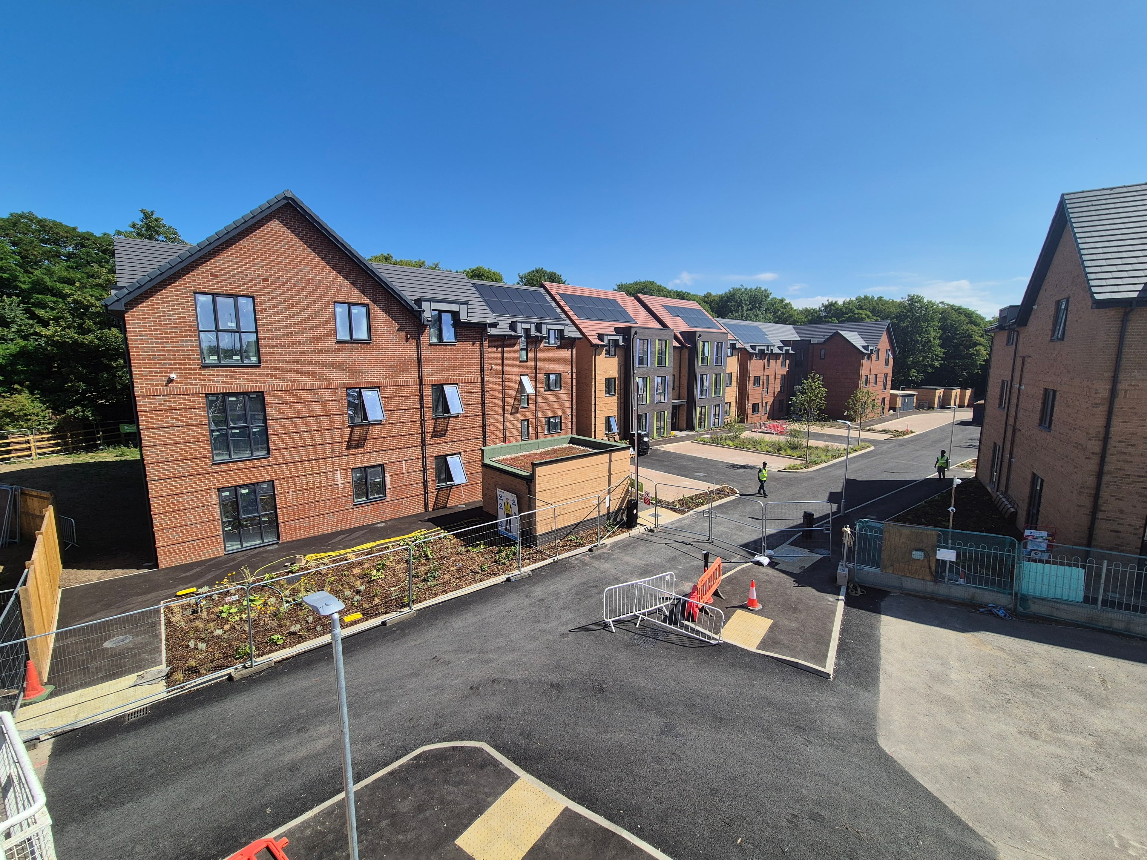 St Peter's (Greenlands and Silverlands), Chertsey: elevated view across the completed development, red-brick blocks with pitched slate roofs and integrated rooftop PV arrays curving along a new internal street under a deep blue sky, mature woodland behind
