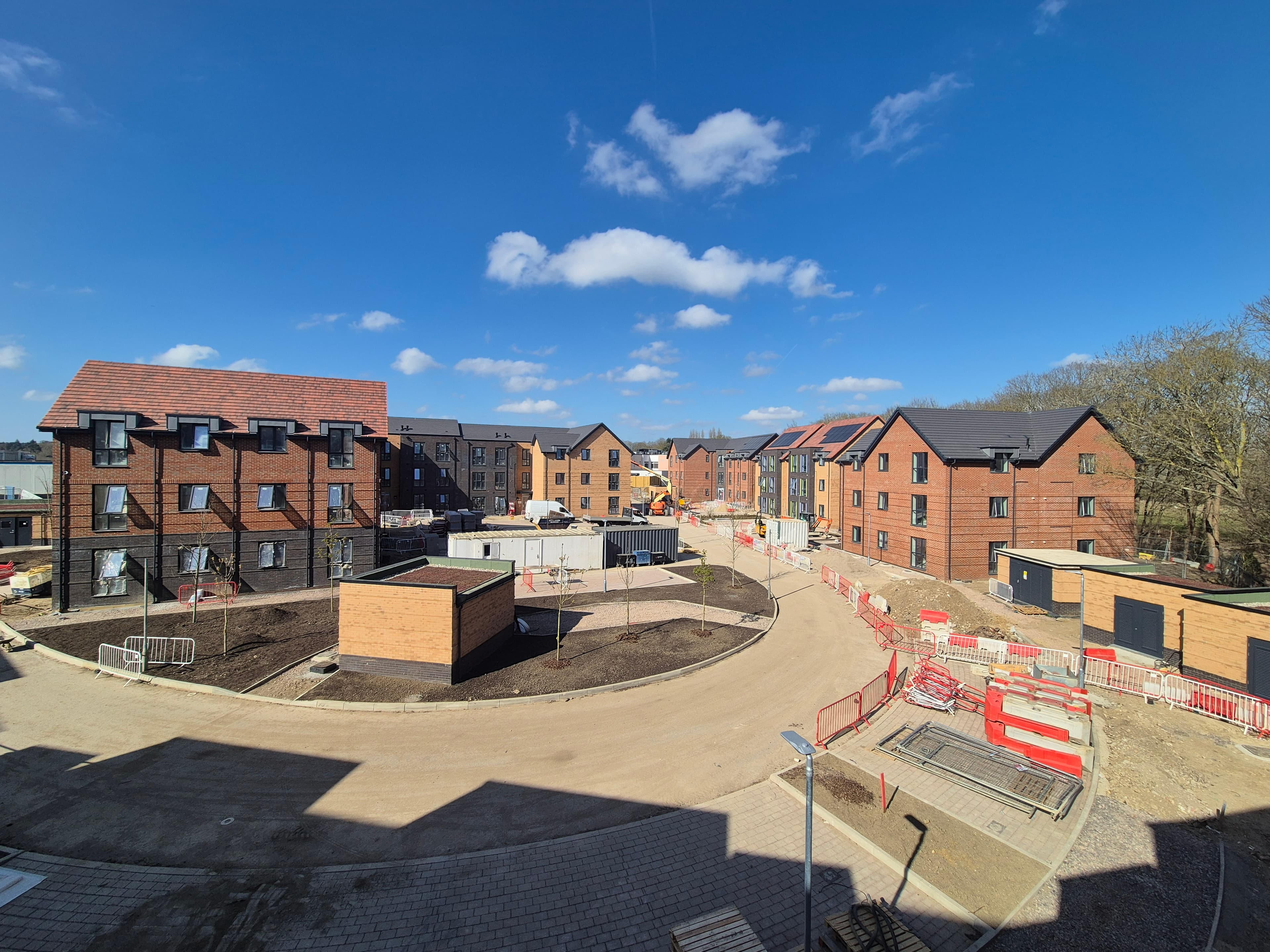 St Peter's (Greenlands and Silverlands), Chertsey: handover-phase view of the completed red-brick blocks with PV-clad slate roofs around a curved tarmac access road, residual Heras fencing in the foreground and woodland beyond