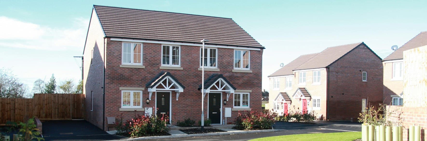Stonehills Close, Meir Heath, Staffordshire: completed pair of two-storey red-brick semi-detached homes with pitched grey roofs and timber porch canopies, set on a planted close in front of an adjacent terrace — Bullock-era affordable scheme.