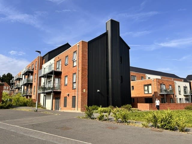 Adlington Road, Sheffield: completed three-storey red-brick and dark-clad apartment block with balconied frontages and a tall dark feature stair tower, set on a wide landscaped boulevard — Sheffield City Council older-persons independent-living scheme.