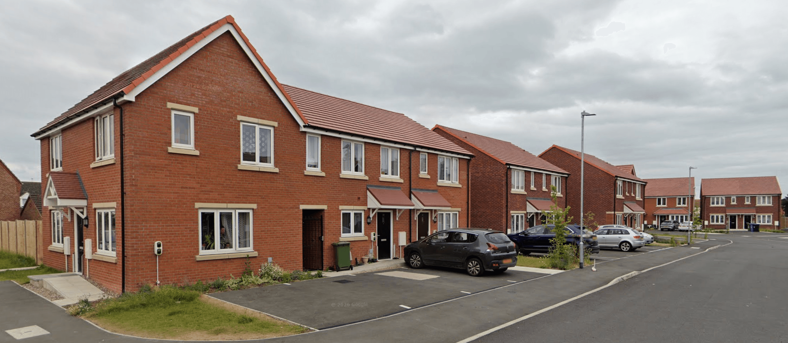 Boston, Lincolnshire, Platform Housing scheme: completed terrace and semi-detached two-storey homes in red brick with terracotta-tiled pitched roofs, white casement windows and shared block-paved drives, viewed along the access road with parked cars on the kerb.