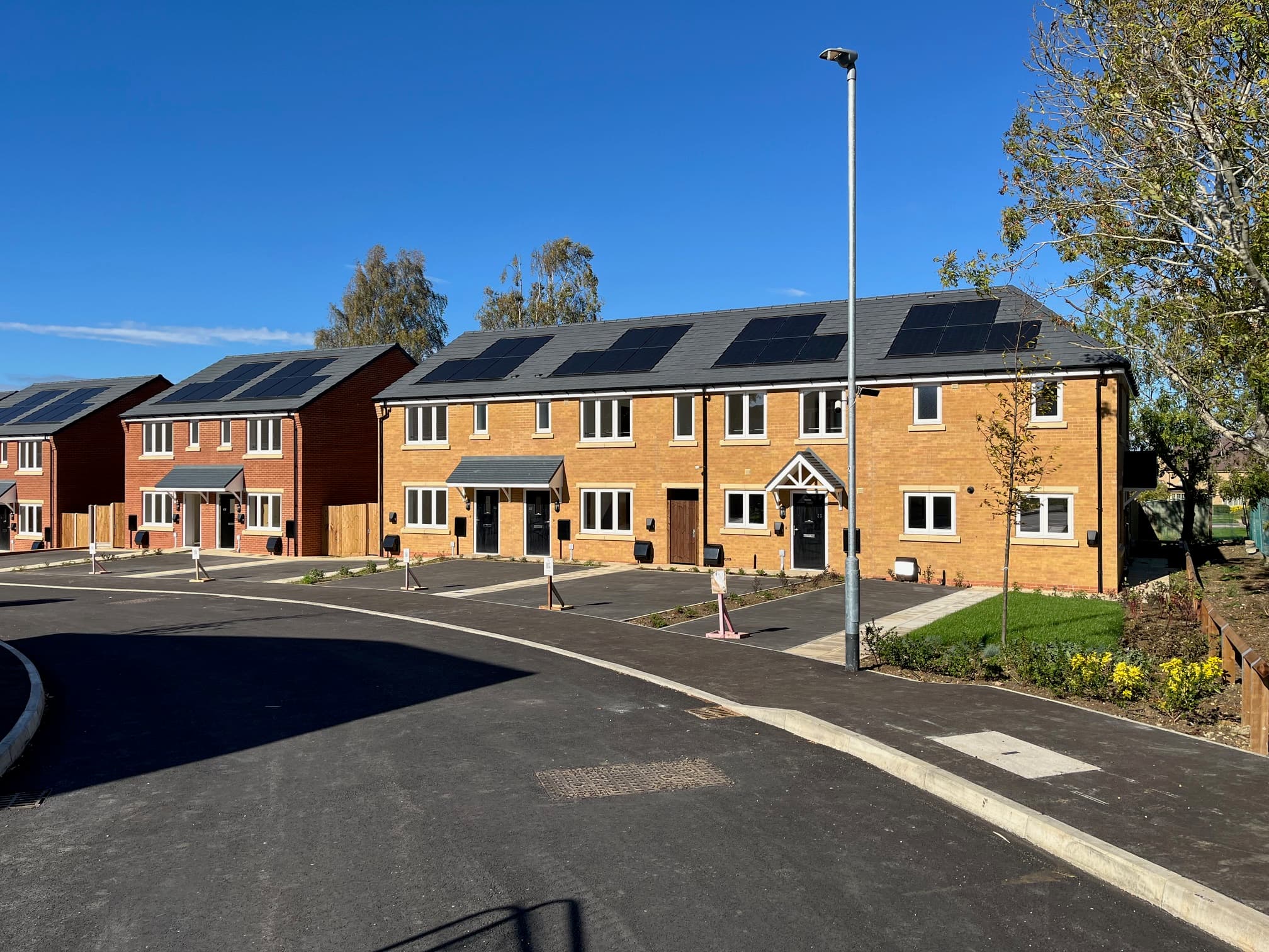 Central Avenue, Northampton: completed terraced and semi-detached homes seen from the new internal road, yellow-brick frontages with darker-brick canopy porches and full-width rooftop PV arrays, autumn afternoon light under a clear blue sky, Futures Housing Group affordable scheme