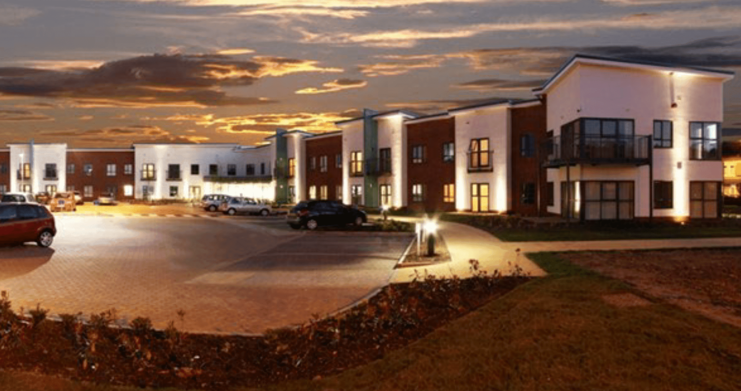 Chestnut Grange, Burton-upon-Trent: completed two-storey extra-care scheme of red brick and white render, set around a curved block-paved entrance drive at dusk, 67 one- and two-bedroom apartments for Trent & Dove Housing, with windows lit and a golden sunset sky behind.