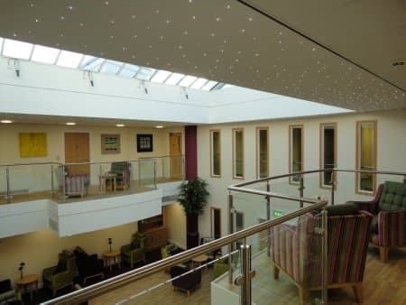 Chestnut Grange interior: bright double-height atrium common-room with roof skylights, white balustraded gallery to the upper floor, striped lounge armchairs and house plants, the social heart of the extra-care home.