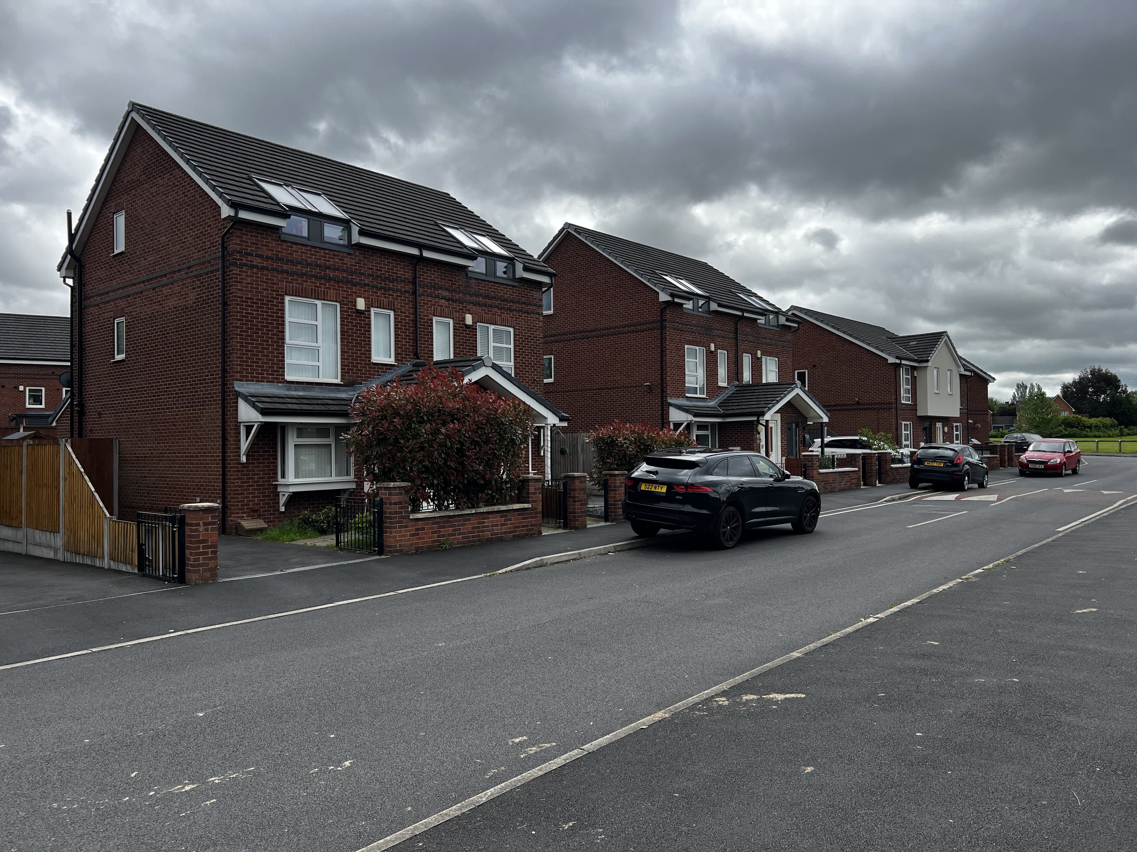 Cotefield Road, Wythenshawe: streetscape of completed two-and-a-half-storey red-brick semi-detached homes with grey pitched roofs, roof-mounted PV panels, and on-street parking on a quiet residential road, Wythenshawe Community Housing Group affordable scheme.