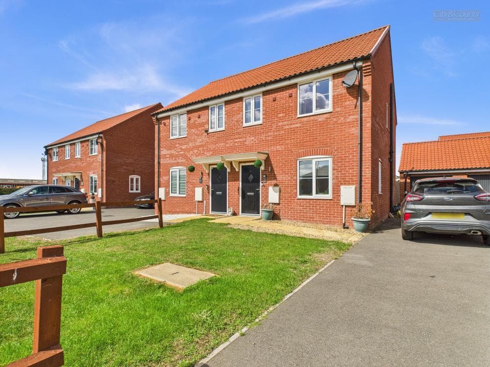 Crease Drove, Peterborough: completed red-brick three-bay semi-detached pair at handover, steep slate pitched roof with hipped ends, brick-arch eyebrow lintels over the windows, dark grey front doors under shallow porch hoods and freshly-laid front lawn, with parked cars and an adjacent terrace visible on either side under a bright blue sky