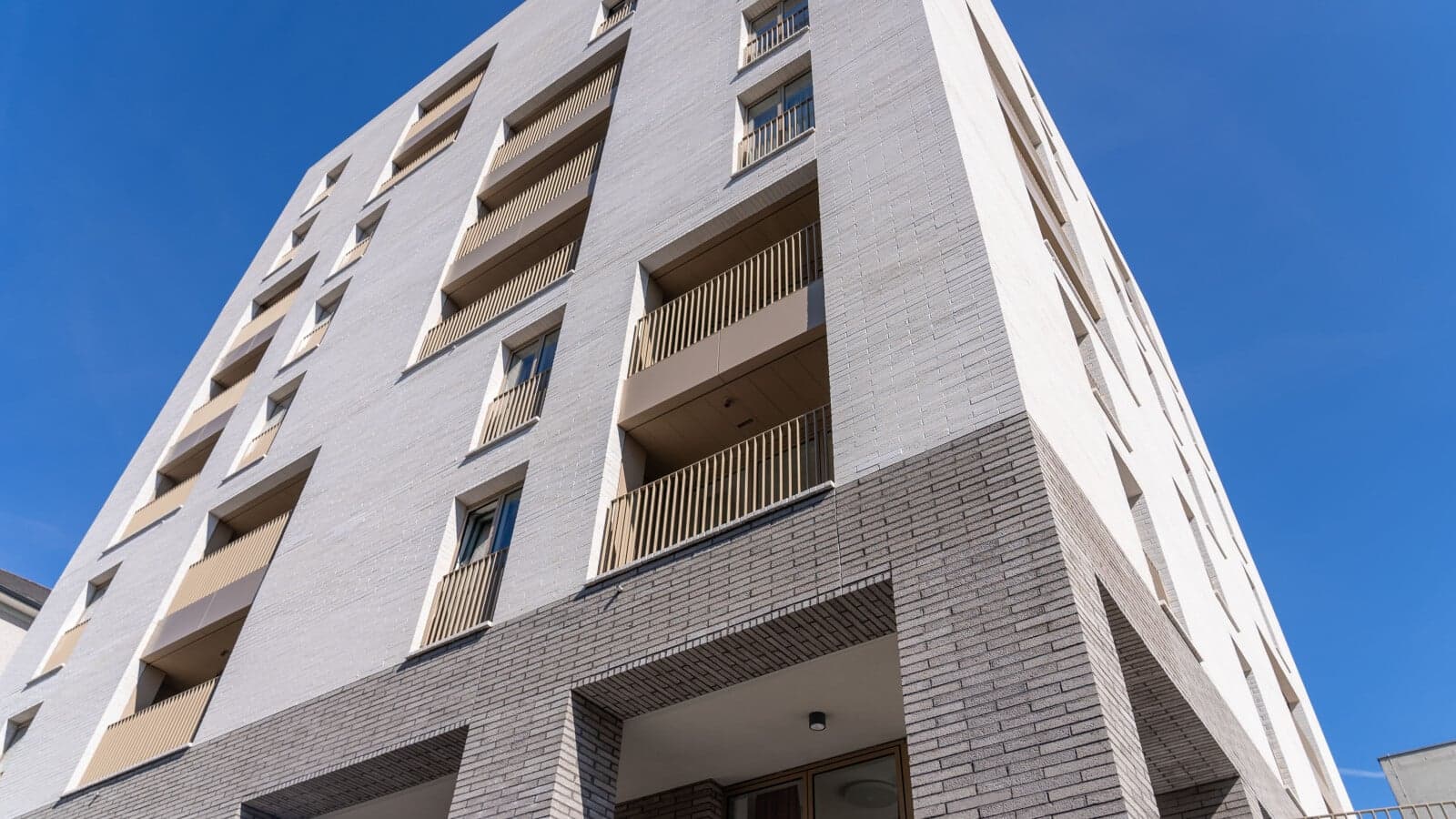 Daventry House (Lisson Arches), Westminster: corner view looking up at the pale-cream brick older-persons block, set-back balconies with timber-screened rails, deep window reveals, ground-floor brick masonry switching to a darker grey-brown band, all against a clear blue sky, Westminster City Council scheme.