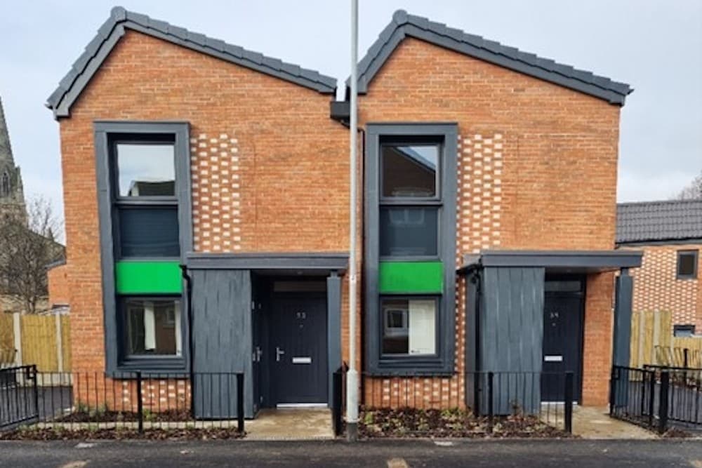 Holbeck Living, Leeds: pair of completed red-brick semi-detached homes with dark grey pitched gables, anthracite window surrounds and patterned brick reveals, church spire visible to the left