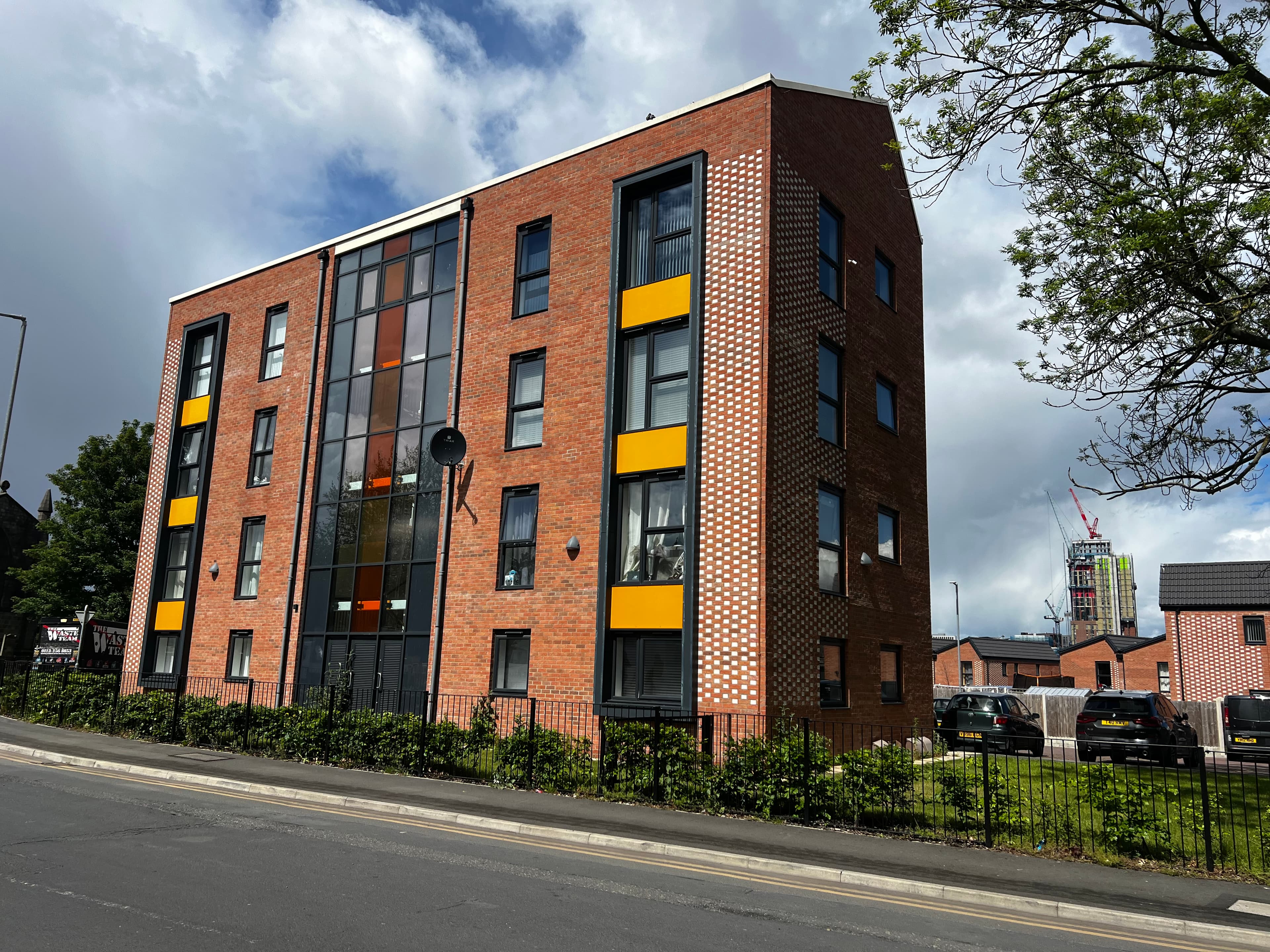 Leeds Meynell, Holbeck: corner view of the three-storey one-bedroom apartment block, red brick walls with yellow accent panels at every window opening, decorative perforated brickwork running the full height of the corner column, modern lift-served entry framed by metal railings.