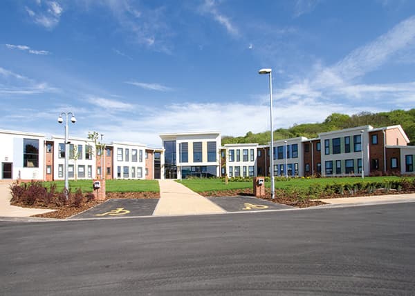 Saltbrook Place, Lye: completed two-storey extra-care blocks with white-rendered upper floors and brick base, central glazed entrance pavilion, lawned forecourt and wooded hillside backdrop, Midland Heart affordable scheme