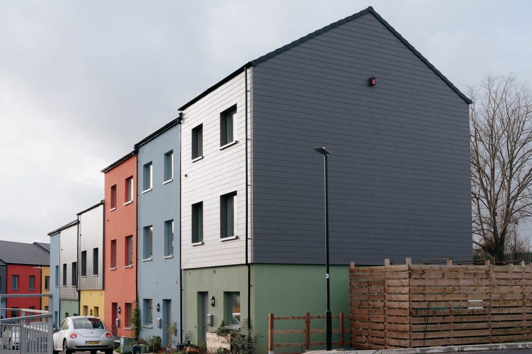 Shaldon Road, Lockleaze (Bristol): completed terrace of modular homes with multi-coloured rendered façades (red, yellow, blue, green, orange) and dark-grey clad gable end, MMC scheme for United Communities and Bristol Community Land Trust