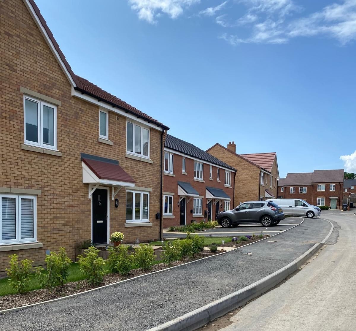 Shepherd's Gate, Shipston-on-Stour: completed row of detached buff-brick houses with grey pitched roofs and tall gables, lining a kerbed street with planted verges and a young hedge, Orbit Group affordable scheme