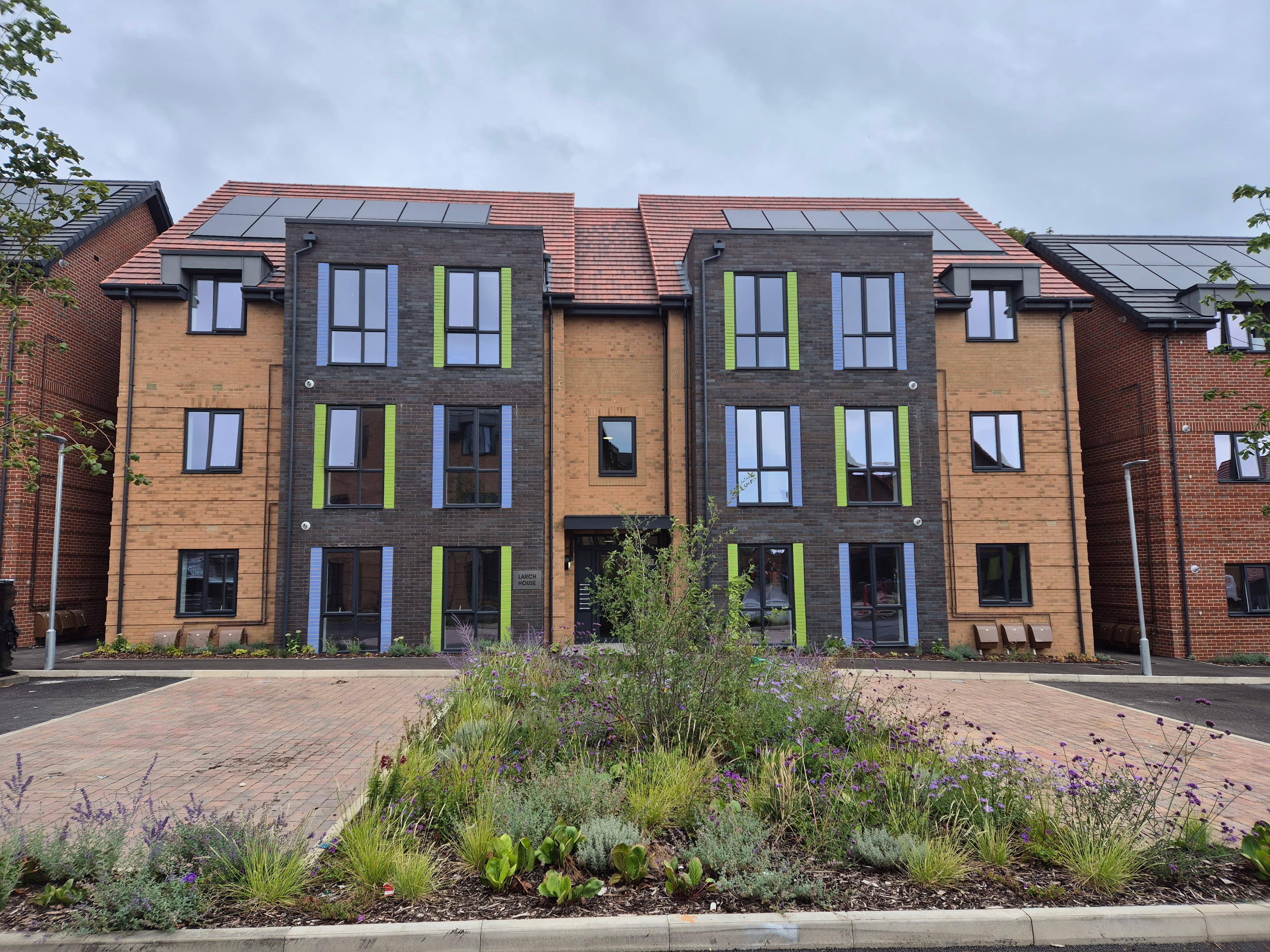 St Peter's (Greenlands and Silverlands), Chertsey: completed three-storey red-brick HAPPI block with twin gables, dark-slate pitched roofs and lime-green window panels punctuating the grey reveal bands, fronted by a wildflower and lavender bed with brick driveway approach, Southern Housing affordable / key-worker scheme