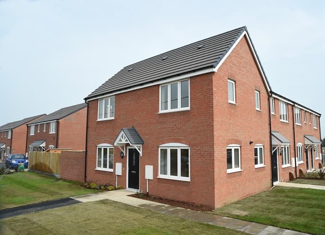 Station Road, Bagworth (Coalville): completed three-bay red-brick semi-detached / end-of-terrace house at handover, slate pitched roof, dark grey window frames and front door under a pitched timber porch canopy, fresh-laid lawn to the front and adjoining brick terraces visible on either side