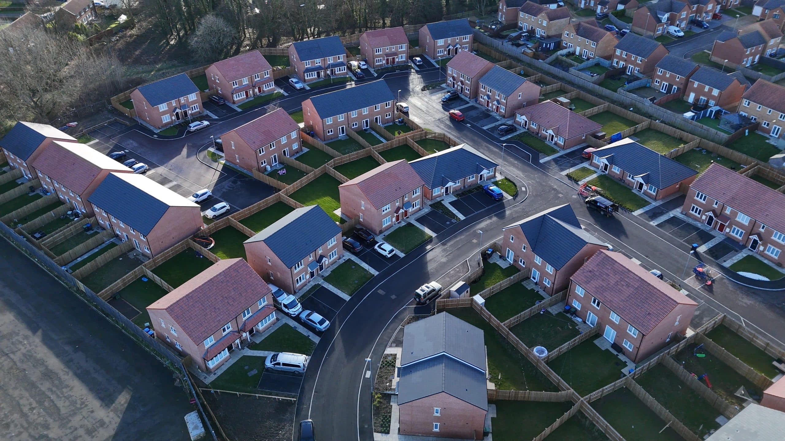 Weavers Close, Peterlee: winter aerial view of the completed estate, clusters of red-brick semi-detached and detached houses with slate pitched roofs arranged around a curved internal street loop, parked cars and freshly-laid landscaping, with the surrounding woodland edge framing the development