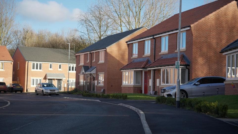 Weavers Close, Peterlee: street-level view of completed red-brick semi-detached houses with slate pitched roofs and grey-framed windows on a bright winter morning, cars parked along the new tarmac estate road, bare trees and woodland behind