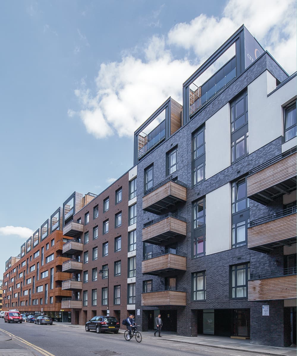 Wharf Road (Wenlock Basin), Islington: street view of the completed six-storey scheme, grey-brick block with timber-clad balconies stepping back at the top, red-brick neighbour and cyclist on the road