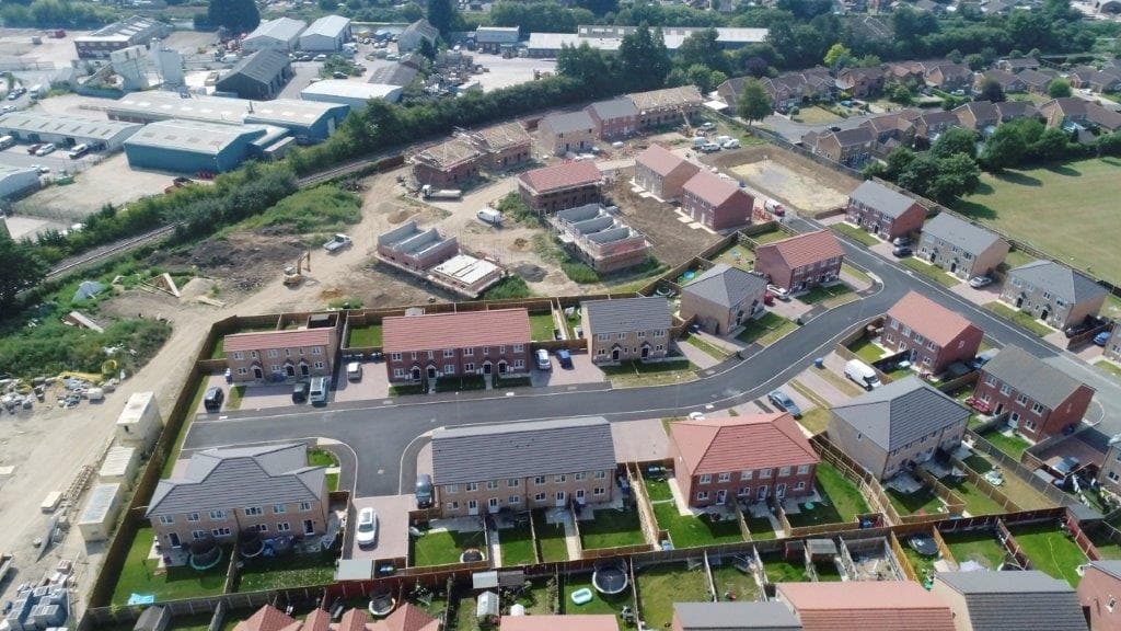 William Morris site, Boston: aerial drone view of the new-build housing scheme mid-construction, completed terraces and semi-detached homes with green gardens in the foreground, plots under construction beyond, neighbouring industrial estate to the rear